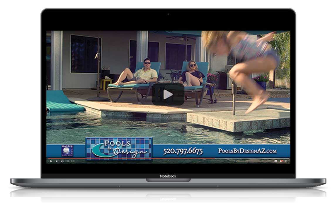 A laptop showing a Pools By Design video on screen paused at the moment a family is lounging around the pool and a kid is doing a cannon ball into the water